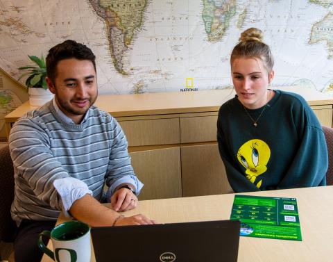 An advisor meeting with a student sitting at a table pointing to a laptop computer