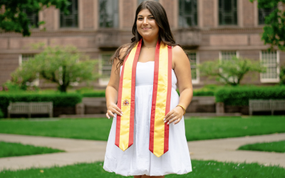 College-aged woman smiling in front of the University of Oregon's Knight Library. She is wearing a Spain flag graduation stole.